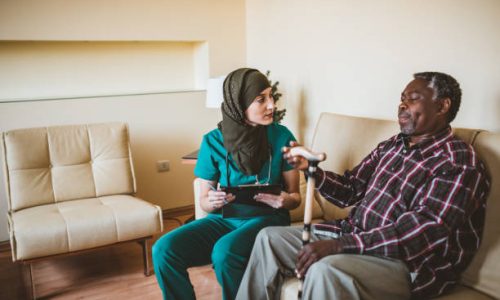 A Muslim female doctor is visiting her older patient of Afro-American ethnicity. An Arabian female healthcare worker is helping her older male patient in the living room at home.